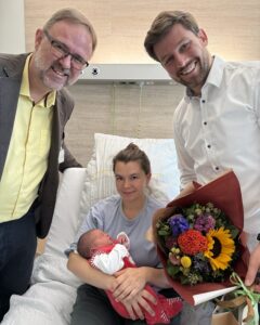 Dr. Matthias Fenski gratuliert Eltern des 1.000 Baby im Vinzenzkrankenhaus 2025. Mutter hält Baby auf dem Arm, Vater daneben mit Blumenstrauß.