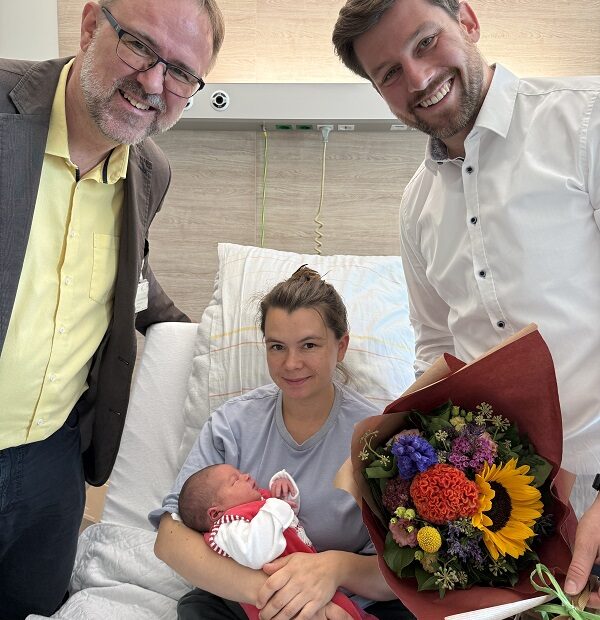 Dr. Matthias Fenski gratuliert Eltern des 1.000 Baby im Vinzenzkrankenhaus 2025. Mutter hält Baby auf dem Arm, Vater daneben mit Blumenstrauß.