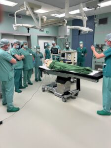 Medizinisches Personal im OP-Saal. Ein Arzt erklärt anhand einer Puppe das Vorgehen bei einem Zwischenfall.