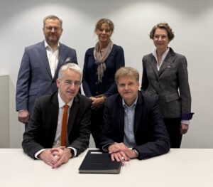 Fünf Personen stehen und sitzen an einem Tisch, auf dem ein Dokument liegt.