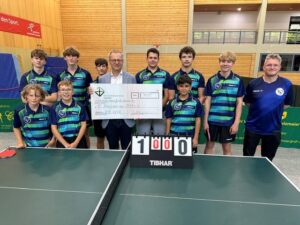 Tischtennisspieler des FC Bennigsen in blau-grünen Trikots hinter einer Tischtennisplatte, in der Mitte Geschäftsführer Schmitt mit symbolischem Scheck